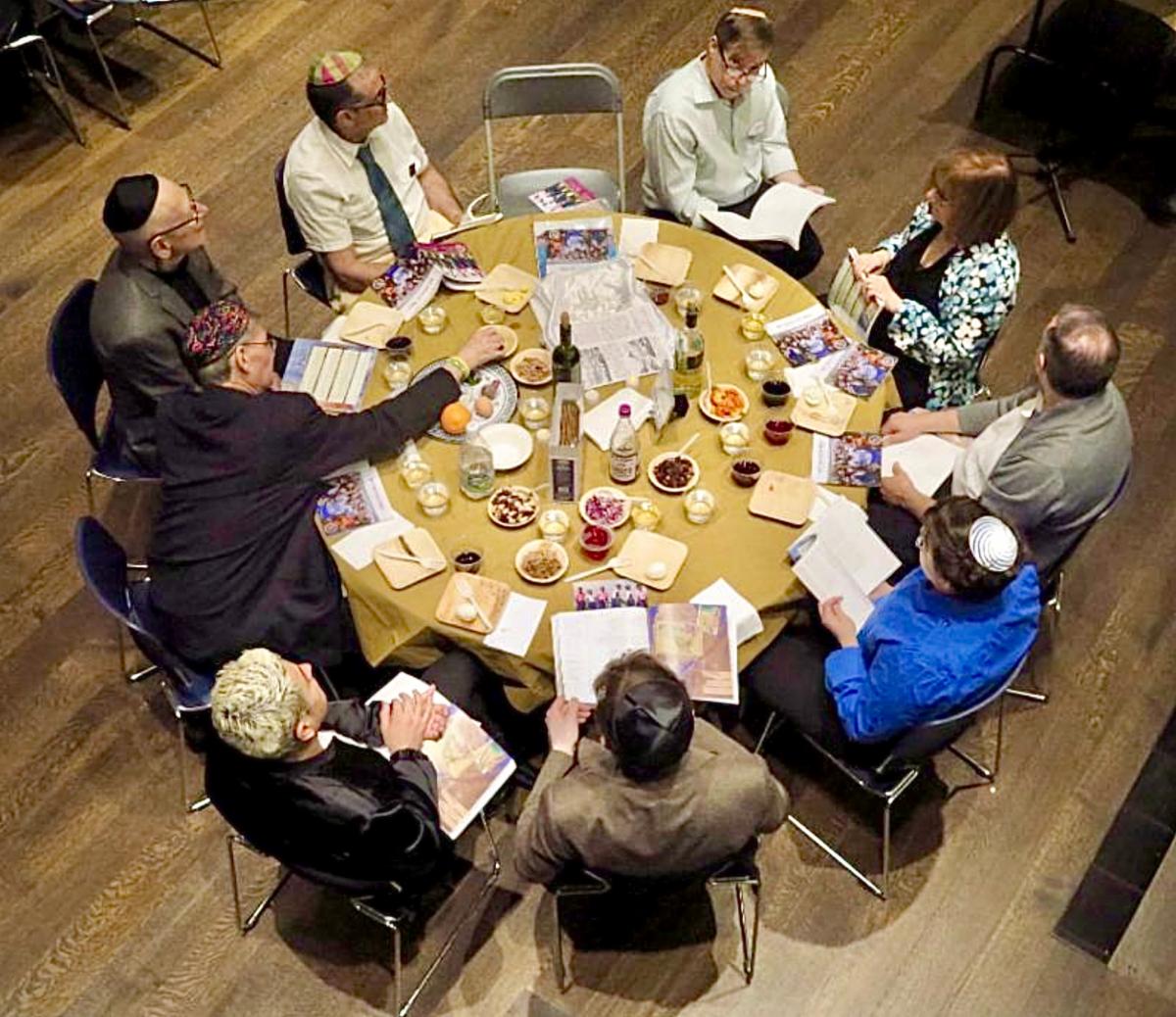 A Seder table in the CBST sanctuary shown from above. Participants sit around the table, which is full of glasses, plates, books, and everything that makes a Seder.