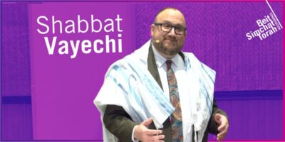 Shabbat Vayechi - Rabbi Klein gestures at the podium, with a purple wash behind him